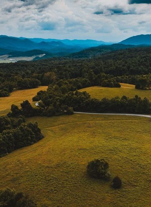 Drone view of wavy roadway between grass meadows with woods behind mounts under cloudy sky